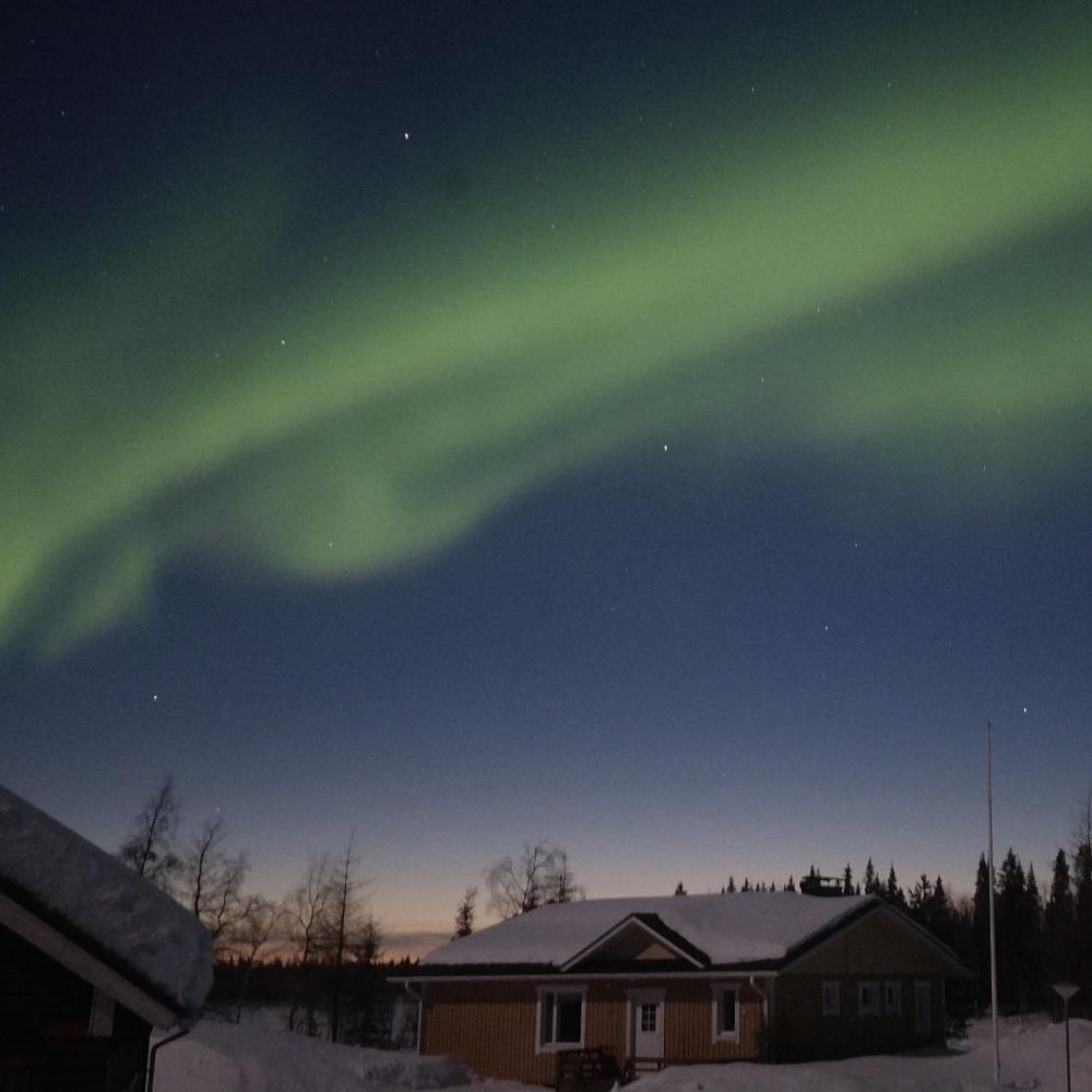Northern lights over a Lapland house at dusk, with the orange glow of a recent sunset still visible on the horizon in early April.