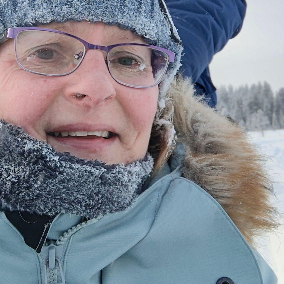 Close-up portrait in Finnish Lapland in extreme cold, with frost crystals covering a knitted hat, scarf and eyelashes, and ice beginning to form at the edges of the glasses.