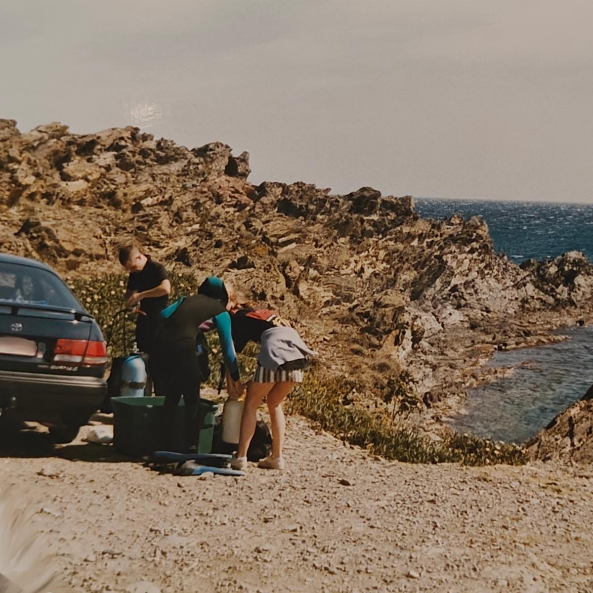 A car parked on rough rocks high above a blue cove, with diving gear and tanks spread out beside it.