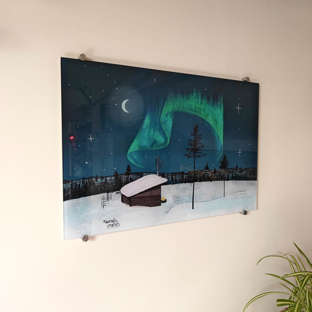 Colour acrylic glass print of a red cabin under a green aurora and crescent moon, mounted with steel standoffs on a light wall beside a houseplant.