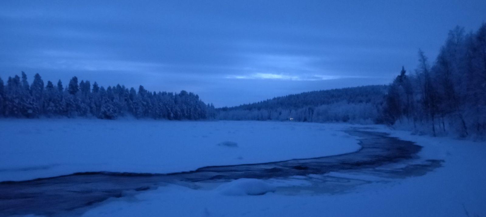 A frozen Lapland river at blue hour in mid-December, a dark open channel of water winding through the snow and ice, forest and hills beyond.