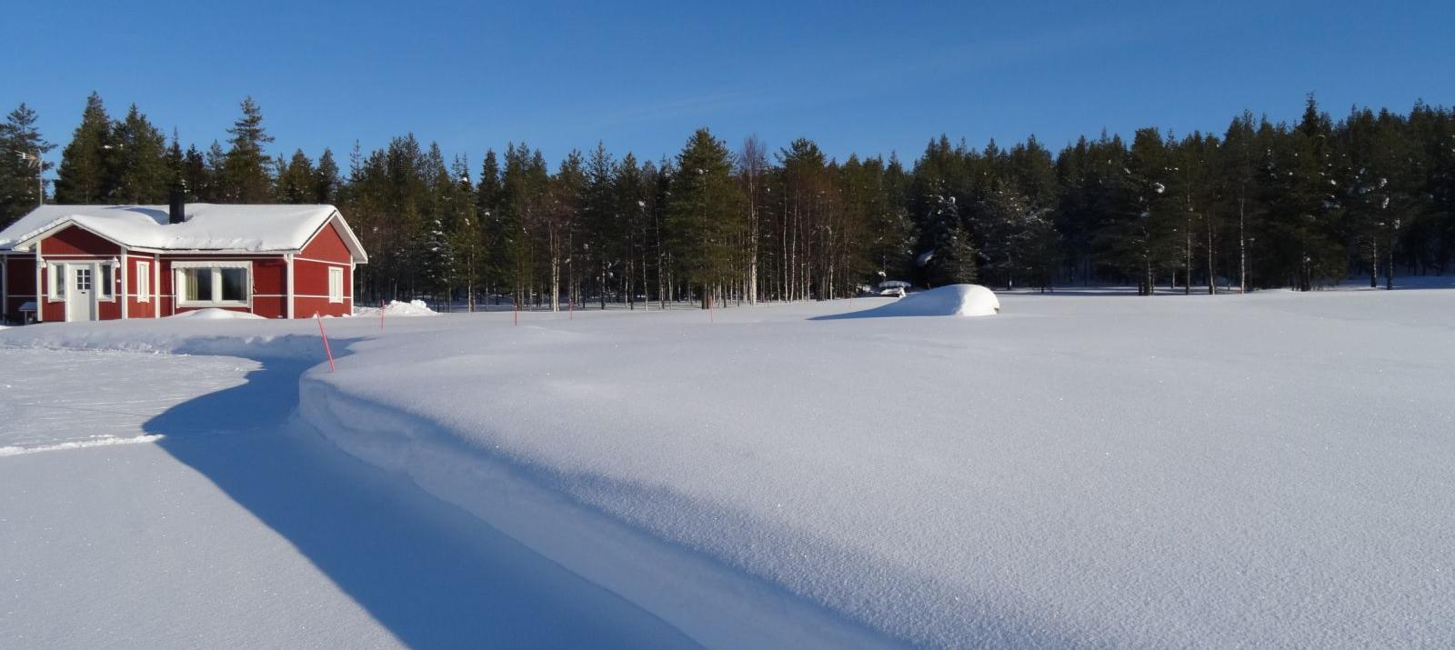 A red Lapland cottage under a deep blue March sky, surrounded by a thick, unbroken snow crust with a long shadow cast across the sunlit snow.