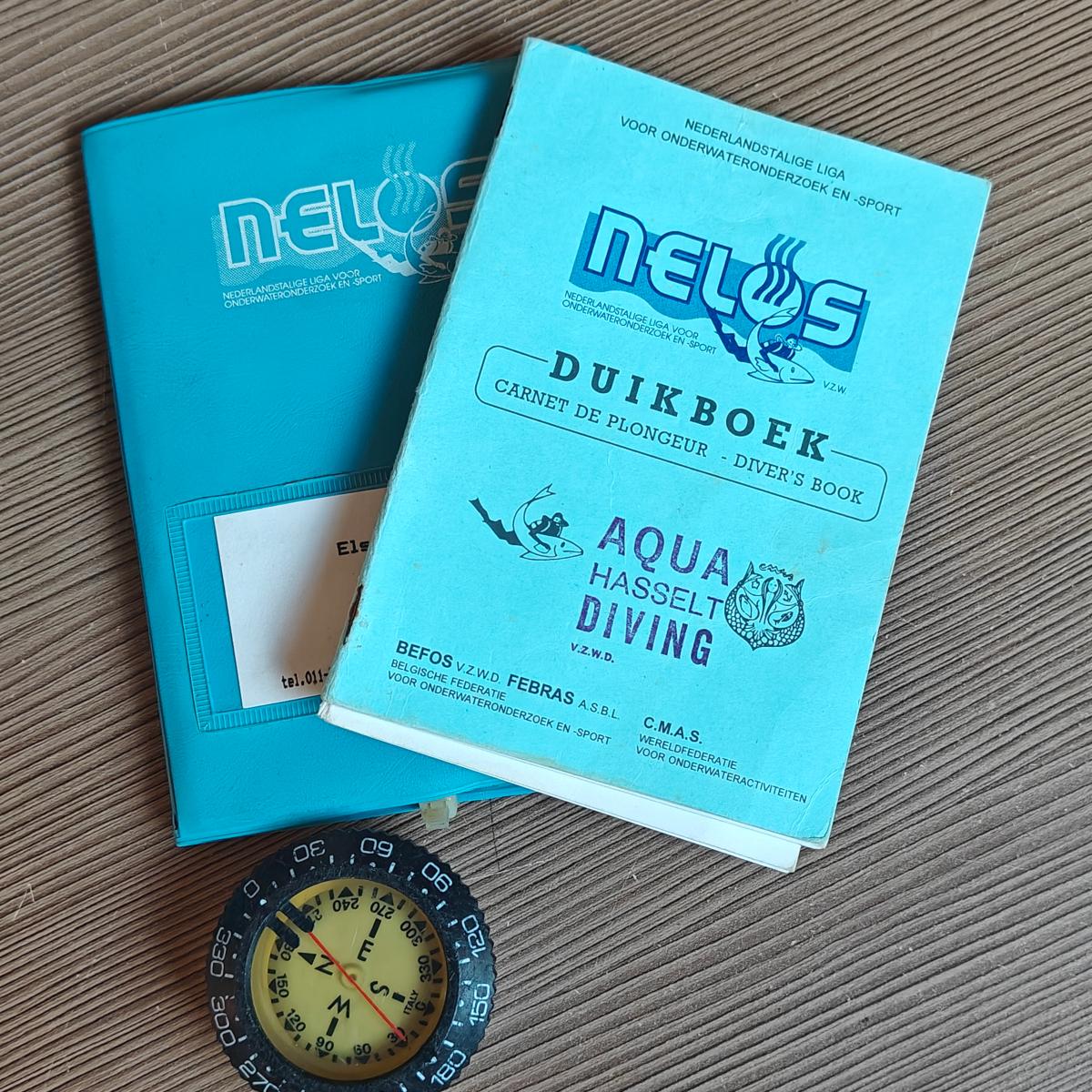 Two pale blue NELOS diver's books on a wooden table, one stamped Aqua Hasselt Diving, next to a black diving compass.