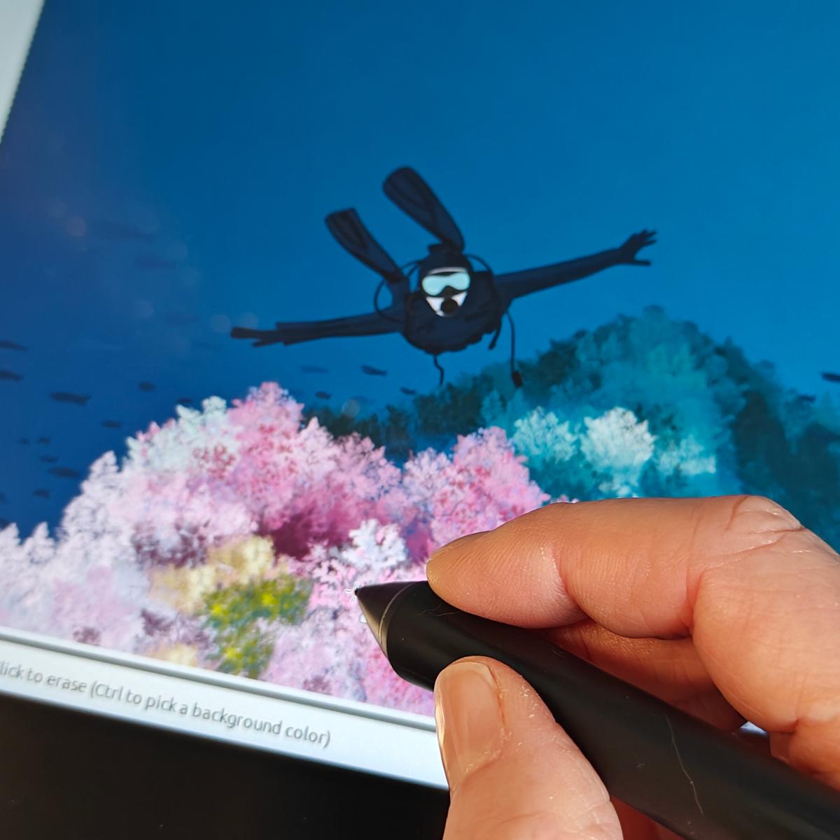 My hand with a stylus working on a drawing tablet drawing an illustration of a diver above pink coral.