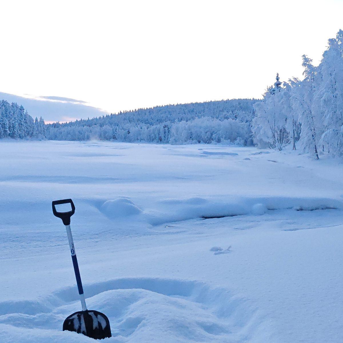 A snow shovel standing in deep snow beside a frozen river in Lapland, the forest heavy with frost under a pale winter sky.