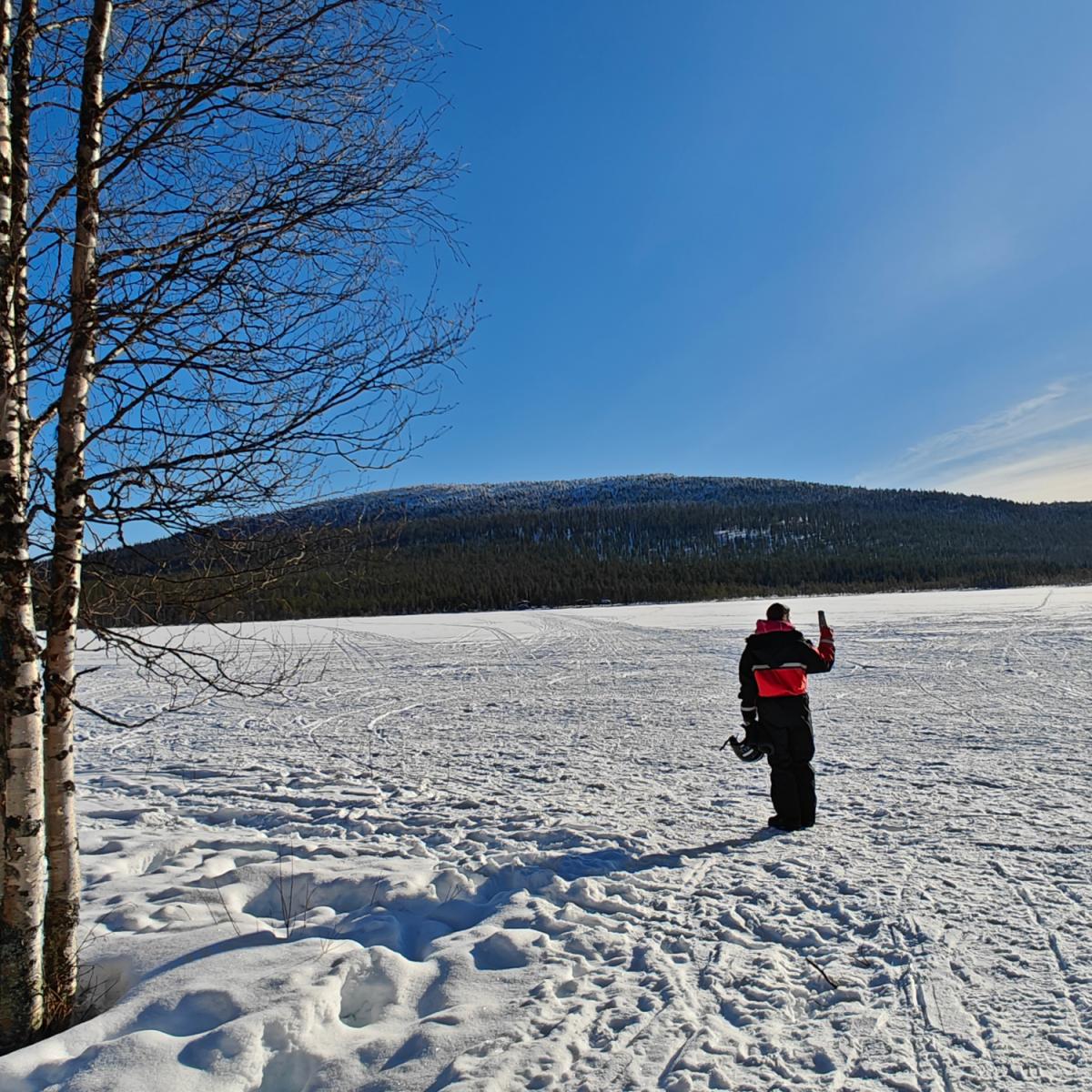 Woman in a black snowmobile suit standing next to her snowmobile on a forest trail near Levi, Finnish Lapland. Aurealis Creatief.