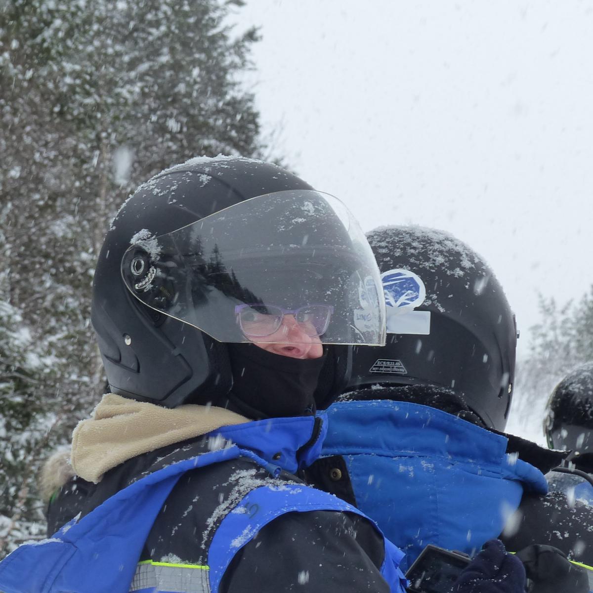 Two snowmobile passengers in blue thermal suits and helmets on a snowy trail in Malangen, Northern Norway, on a grey spring day. Aurealis Creatief.